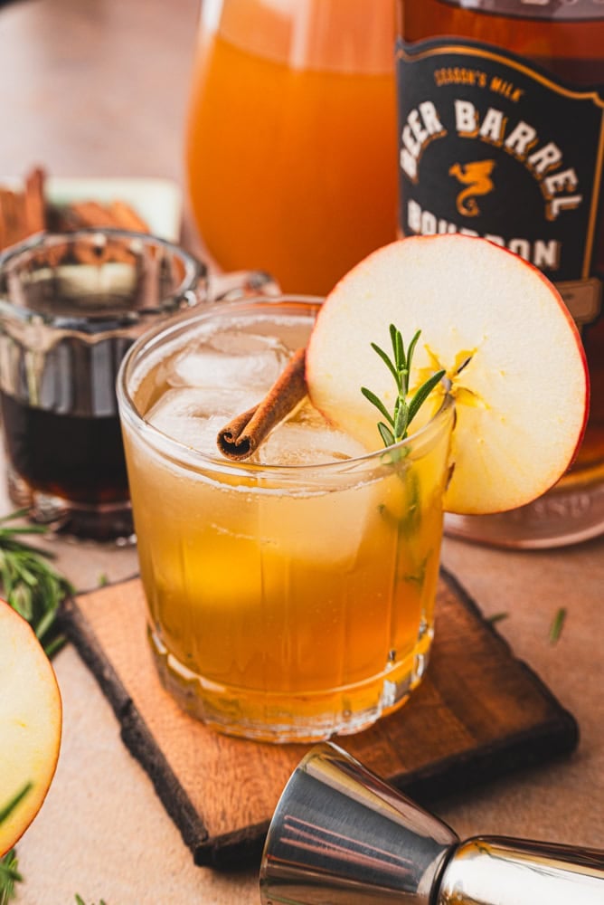 apple cider bourbon cocktail with apples and a little jar of maple syrup in the background. The top is garnished with a round apple slice, cinnamon stick, and rosemary.