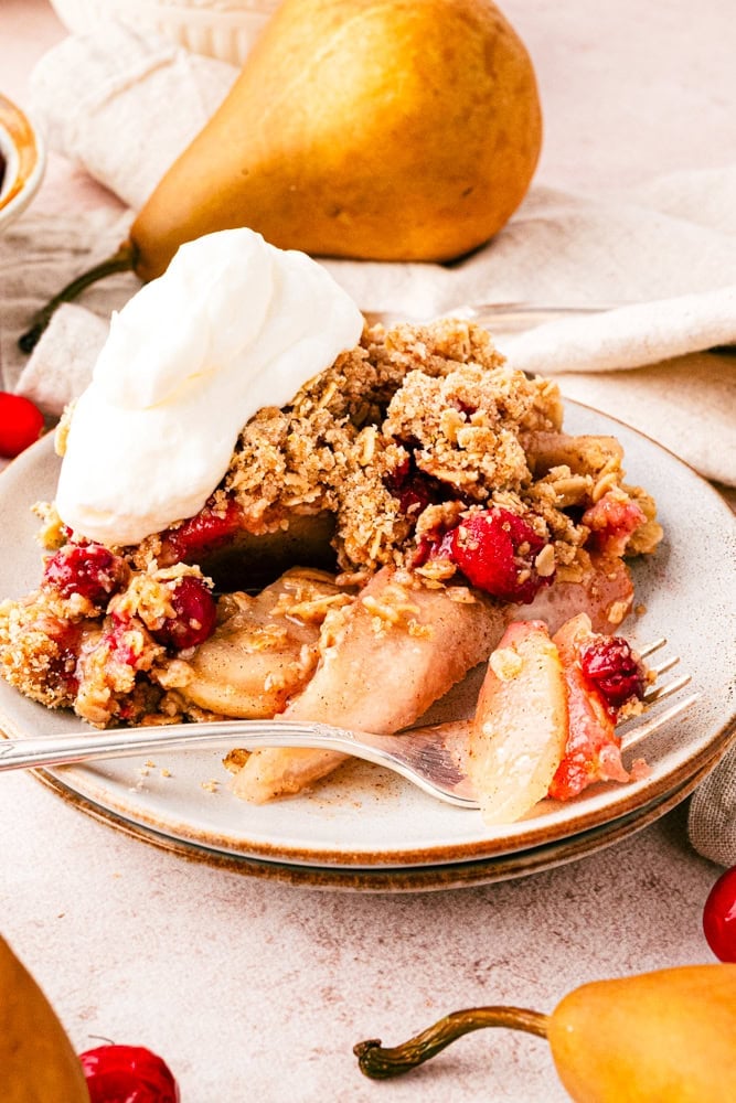 side view of pear crisp on a plate with a fork and a dollop of whipped cream. there's a pear in the background