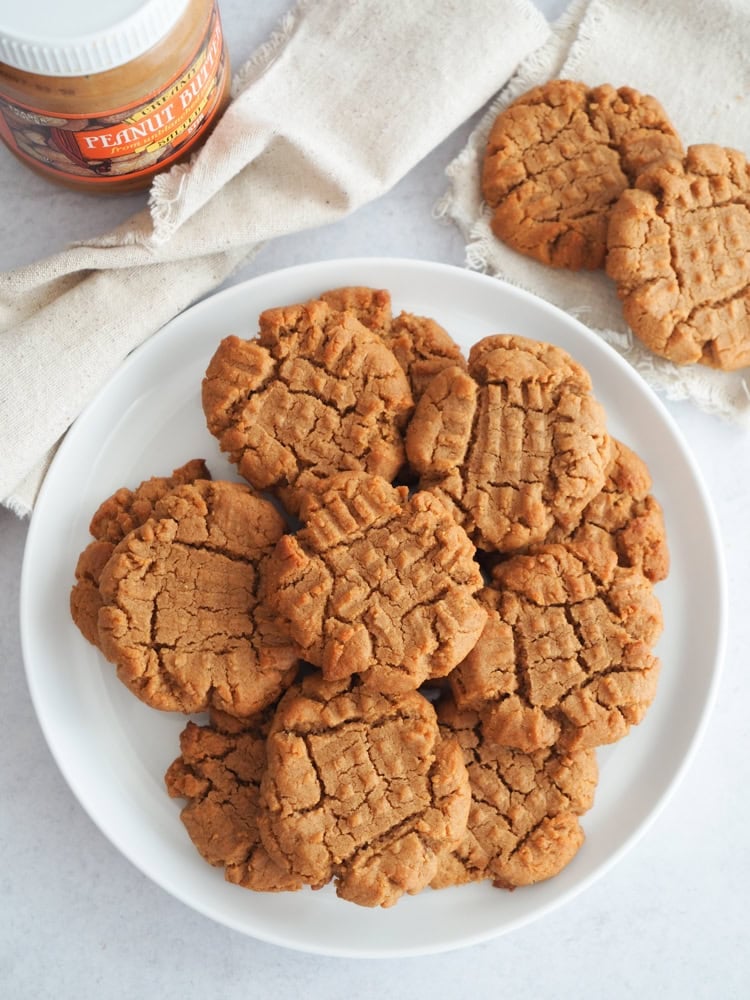 white plate with 4 ingredient peanut butter cookies. there's a jar of peanut butter to the left and two extra cookies to the right.