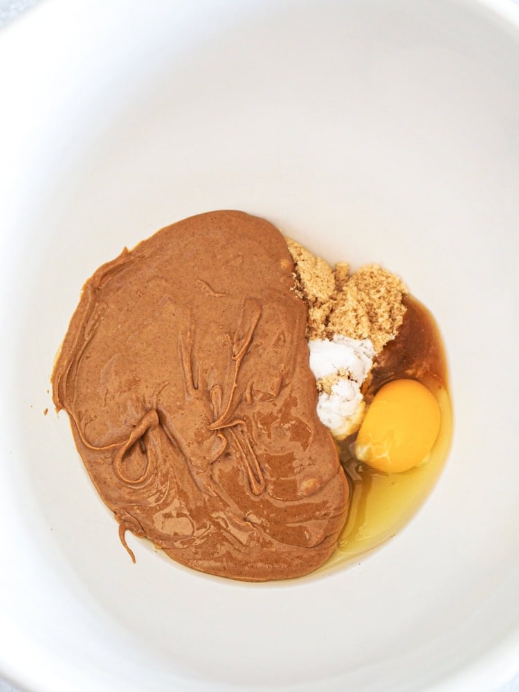 close up of 4 ingredients for peanut butter cookies in a white bowl: peanut butter, egg, light brown sugar, baking soda
