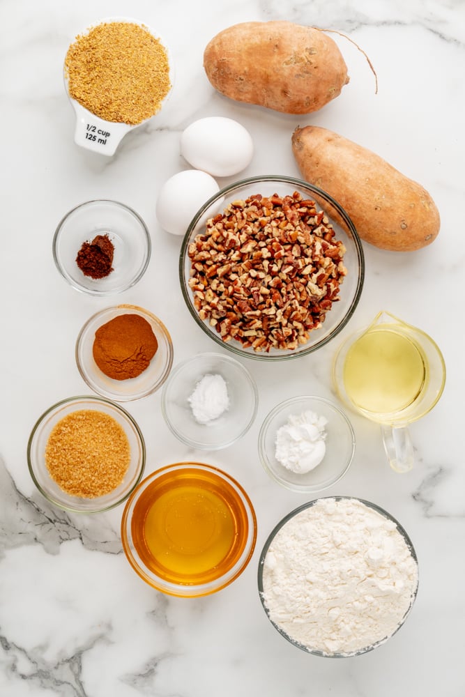 overhead shot of ingredients to make sweet potato muffins, with everything measured out in bowls, and sweet potatoes and eggs to the side