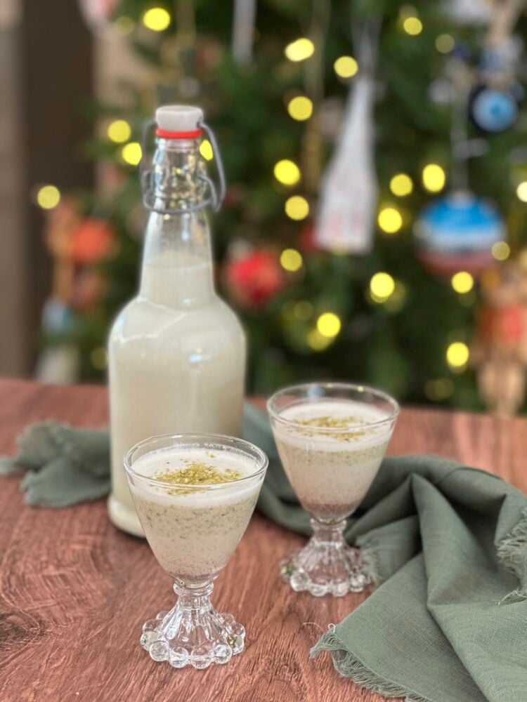 Bottle of Pistachio coquito with two small serving glasses. There's a Christmas tree in the background.