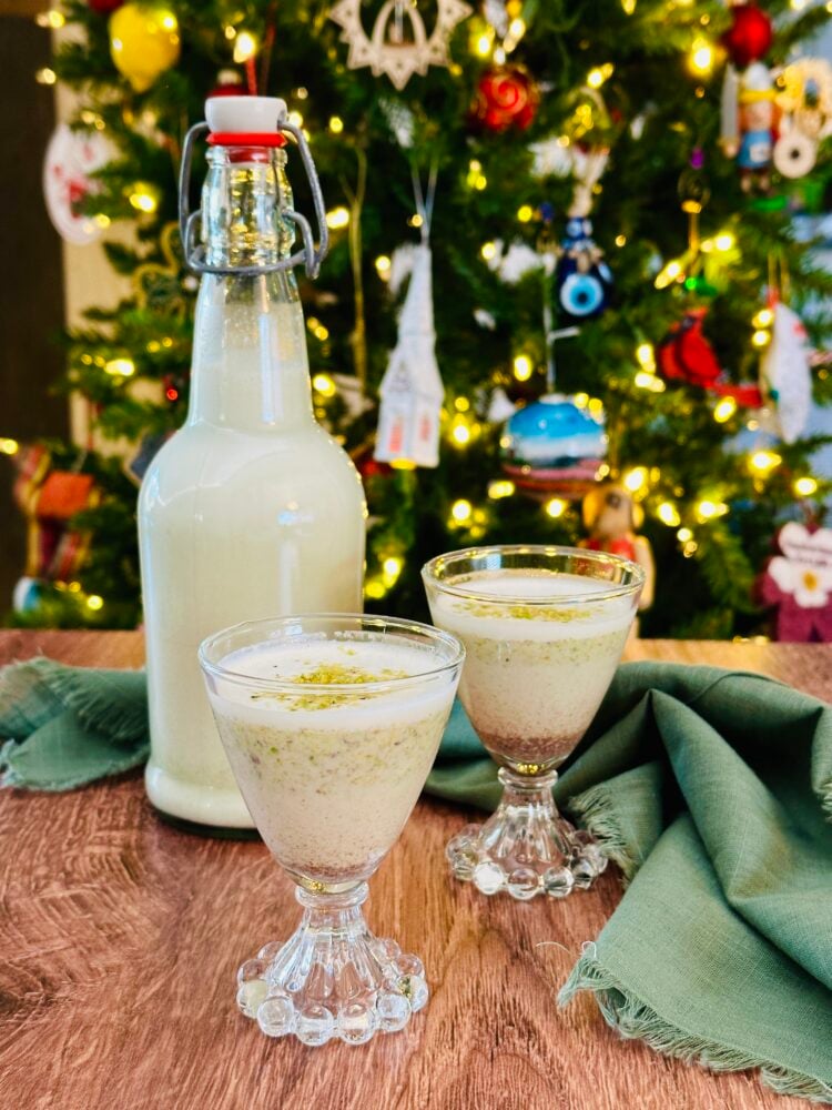 Pistachio coquito in a bottle with two serving glasses and a Christmas tree in the background.