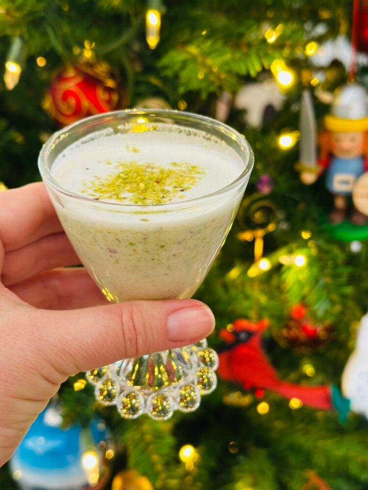 Someone holding a glass of Pistachio coquito next to a Christmas tree.