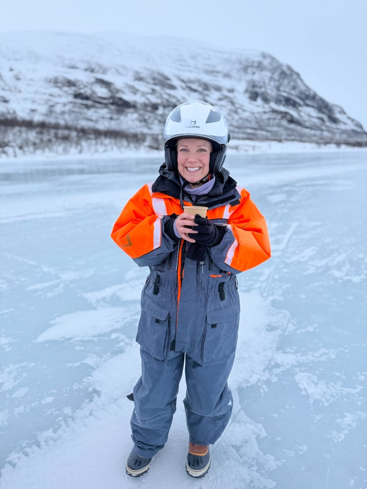 Rachelle wearing a snowsuit holding a hot cup of lingonberry juice while standing on a frozen lake