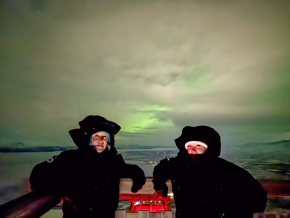Rachelle and friend in black snowsuits looking out at Abisko from the top of a mountain