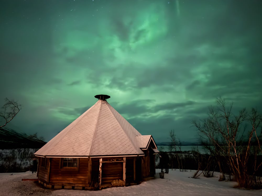 Hut at Abisko tourist station with light green aurora in the clouds behind it.