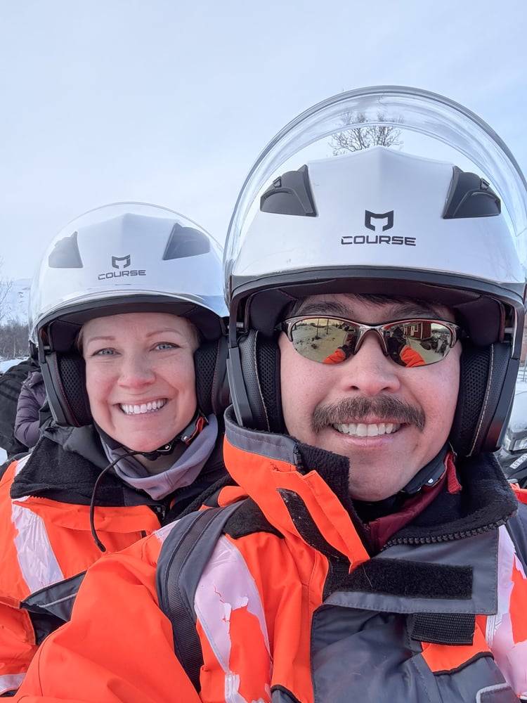 Selfie of Rachelle and Pete on a snowmobile. They're wearing a bright orange snow suite and helmets