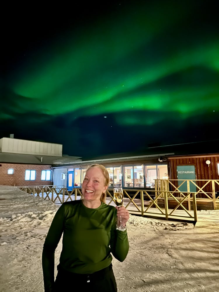Rachelle holding a glass of champagne outside of Abisko restaurant with the northern lights above in the sky.