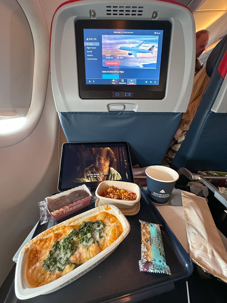 window seat in Delta Comfort Plus with meal including pasta, tiramisu, salad, and a cup of water. There's also an iPad showing extra space.