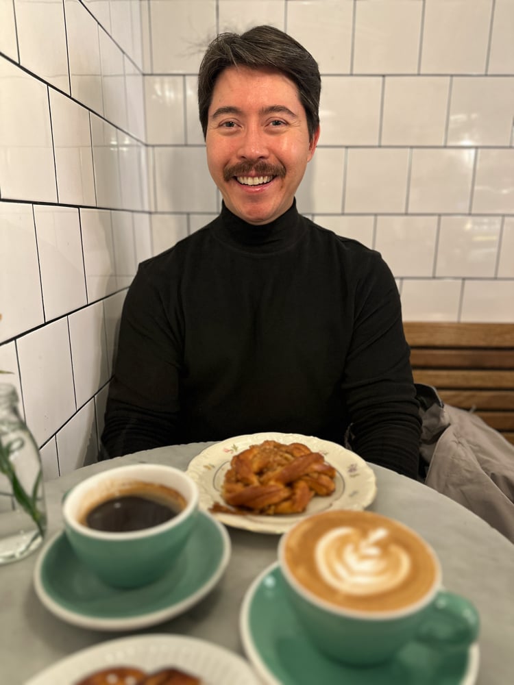 Pete wearing a black turtleneck at a cafe. There's coffee and a cinnamon bun on the table.