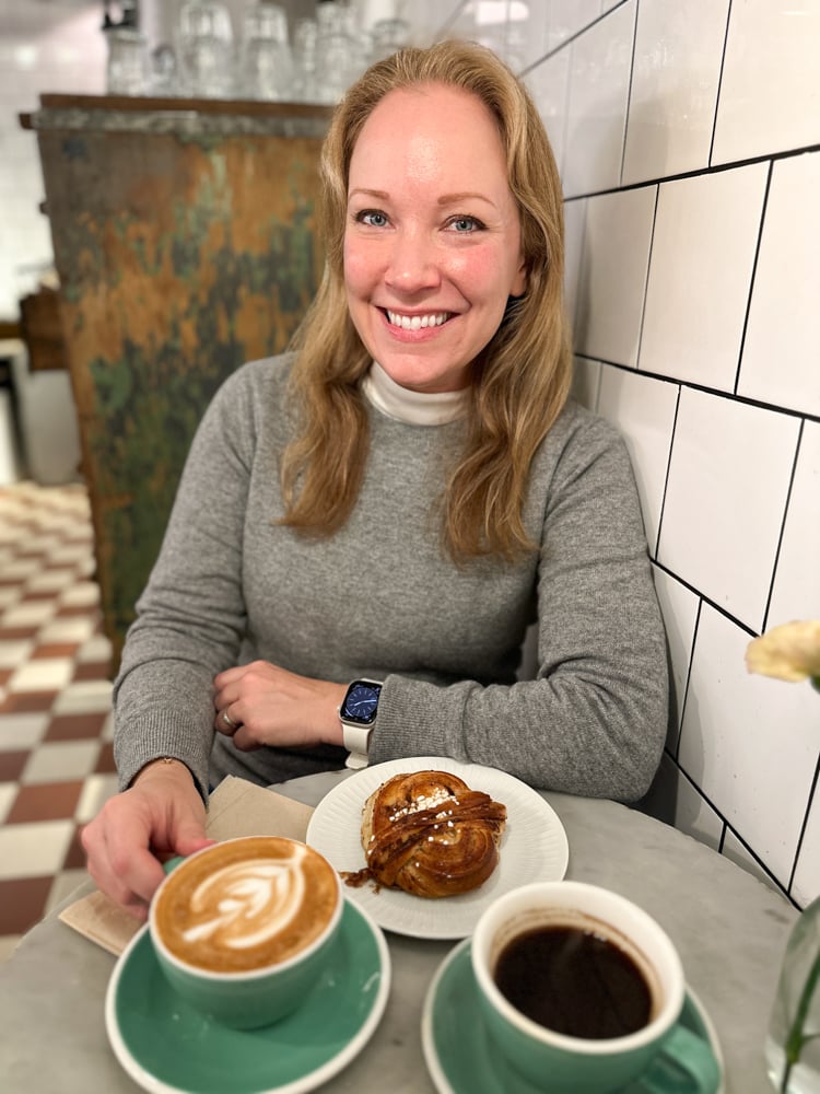 Rachelle wearing a grey sweater in a cafe. There's coffee and a cinnamon bun on the table.