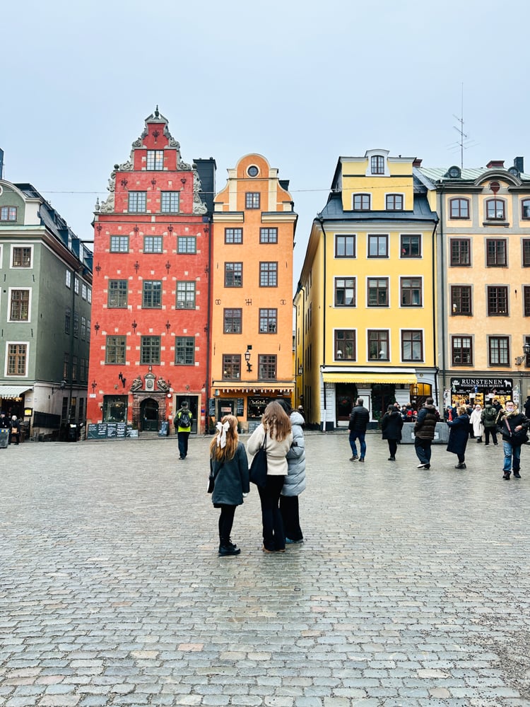 Orange and yellow historic buildings in a square in Stockholm with three girls looking up at them. One girl as a white bow in her hair.