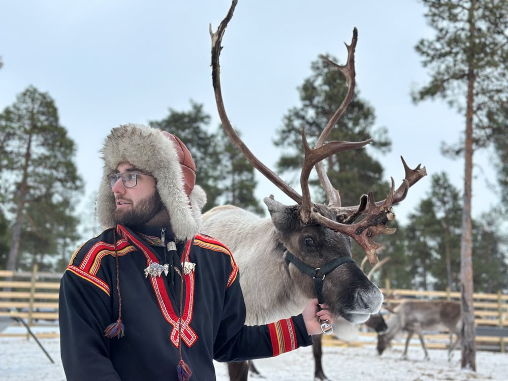 Sami guide in traditional outfit with red trim, holding the reins to a reindeer with bit antlers.
