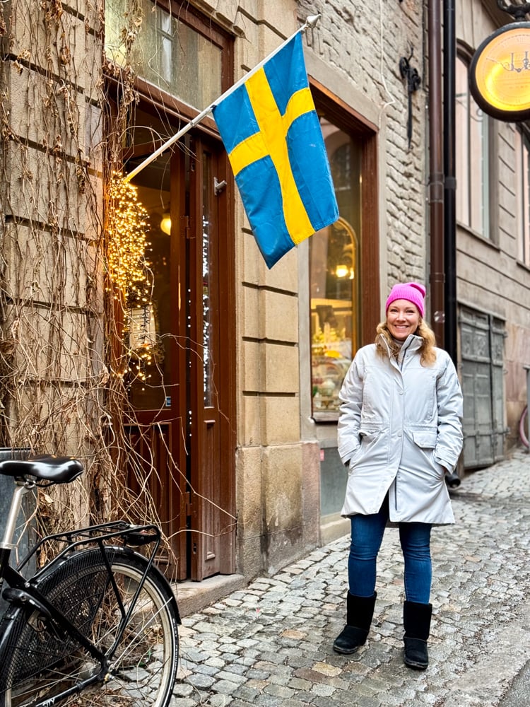 Rachelle wearing a pink hat and white parka on a stone street next to a historic building with a Swedish flag.