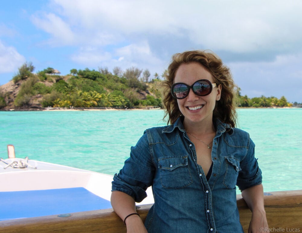 Rachelle wearing a jean shirt overtop a bathing suit, and big sun glasses on the back of a boat in Bora Bora with turquoise water behind her