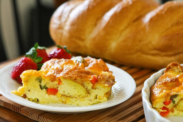 slice of Italian breakfast strata on a white plate with strawberries as garnish on the side and a loaf of Italian bread in the background