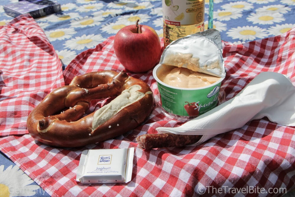 Picnic spread on a red and white checkered cloth with an apple, pretzel, spreadable cheese, sausage, and a square of chocolate.