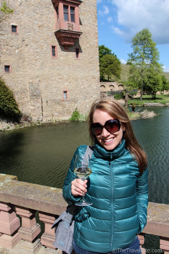 Rachelle in a teal jacket posing in front of the castle-like tower at Schloss Vollrads with a bottle of Riesling in hand.