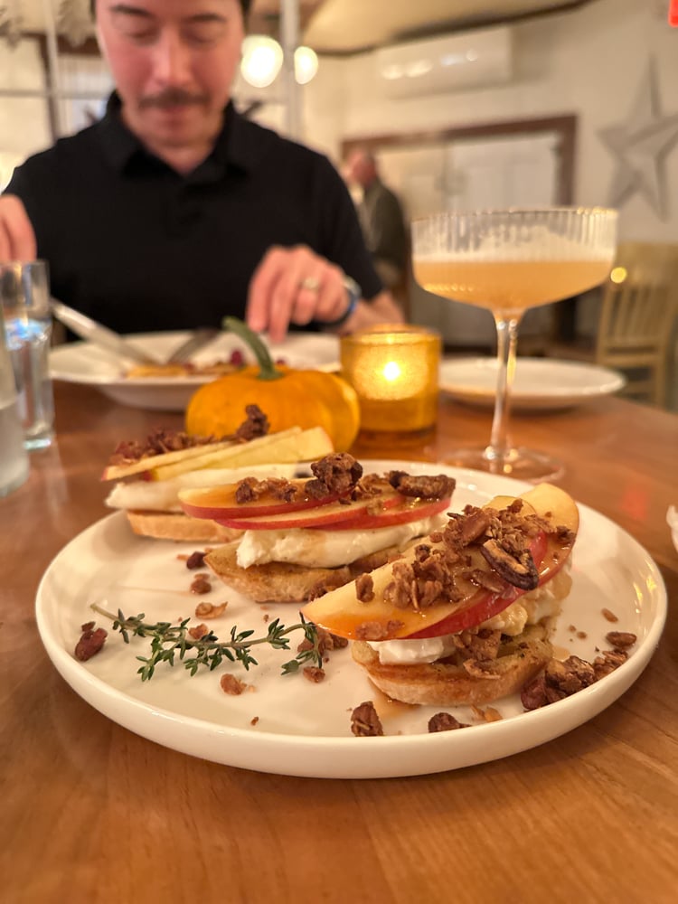 Pete blurred in the background, with autumn apple covered toast, a mini pumpkin, and a cocktail in the foreground