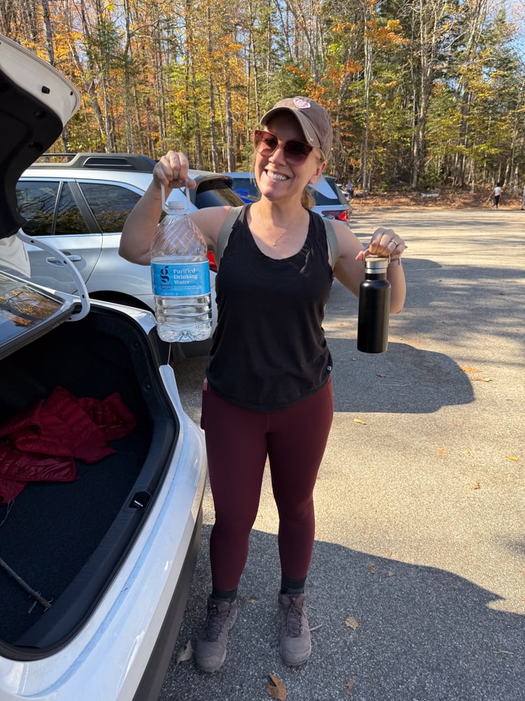 Rachelle standing by the car trunk holding a gallon of water and her hiking water bottle